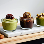 A vibrant and inviting display of various healthy, plant-based sweet treats. Features include a rich avocado chocolate mousse in a glass ramekin, colorful energy balls made with dates and nuts, and a slice of a golden banana-oat cake. Each item showcases natural ingredients and tempting textures. Set on a clean, light-colored kitchen counter or a rustic wooden table, with soft, natural daylight illuminating the scene. A few fresh ingredients like whole dates, almonds, and berries are artfully scattered nearby. Professional food photography, high resolution, soft focus on the delicious textures, appropriate content, safe for work, fully clothed, modest, family-friendly, perfect anatomy, correct proportions, natural pose.